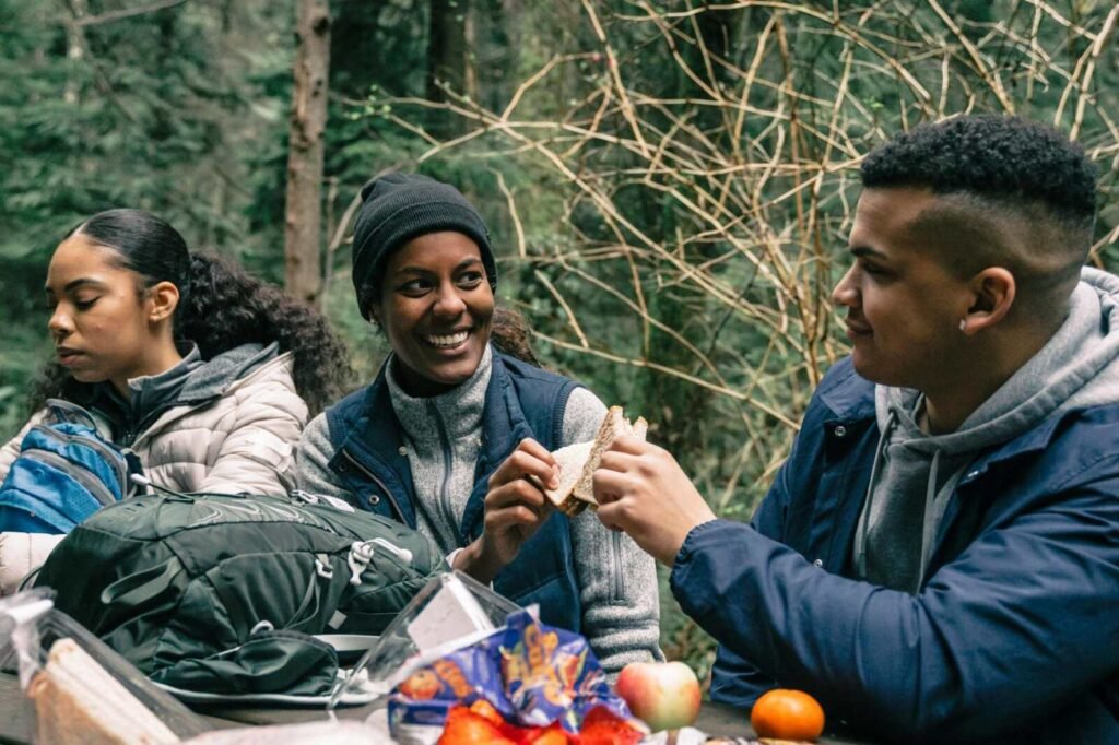 Three friends share a meal while hiking in a forest, enjoying nature and laughter.