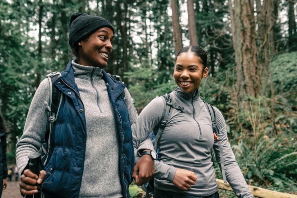Two women hike a forest trail, smiling and enjoying nature's beauty.